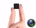 A miniature black square security camera held between two fingers to show its small size, with a circular inset graphic of a larger camera lens in the bottom right corner on a white background.
