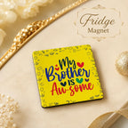 A square yellow fridge magnet with the phrase 'My Brother Is Awesome' in colorful lettering, styled on a beige surface with decorative gold ribbon and holiday ornaments. A label in the top right reads 'Fridge Magnet'.