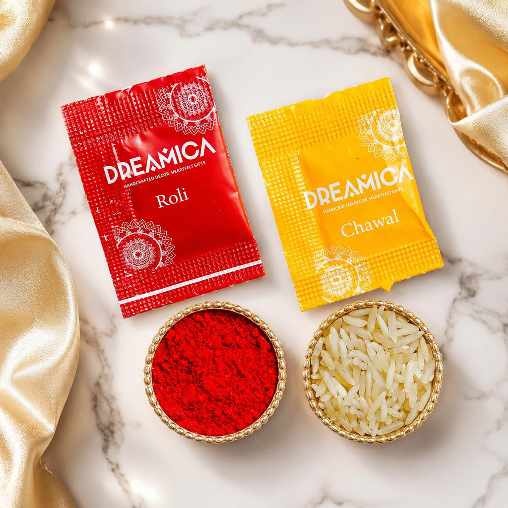 Two Dreamica branded packets, one red labeled Roli and one yellow labeled Chawal, arranged above two small bowls on a white marble surface. One bowl contains bright red powder and the other contains white rice grains, with gold satin fabric draped around the scene.