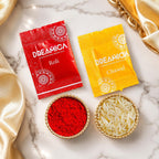 Two Dreamica branded packets, one red labeled Roli and one yellow labeled Chawal, arranged above two small bowls on a white marble surface. One bowl contains bright red powder and the other contains white rice grains, with gold satin fabric draped around the scene.