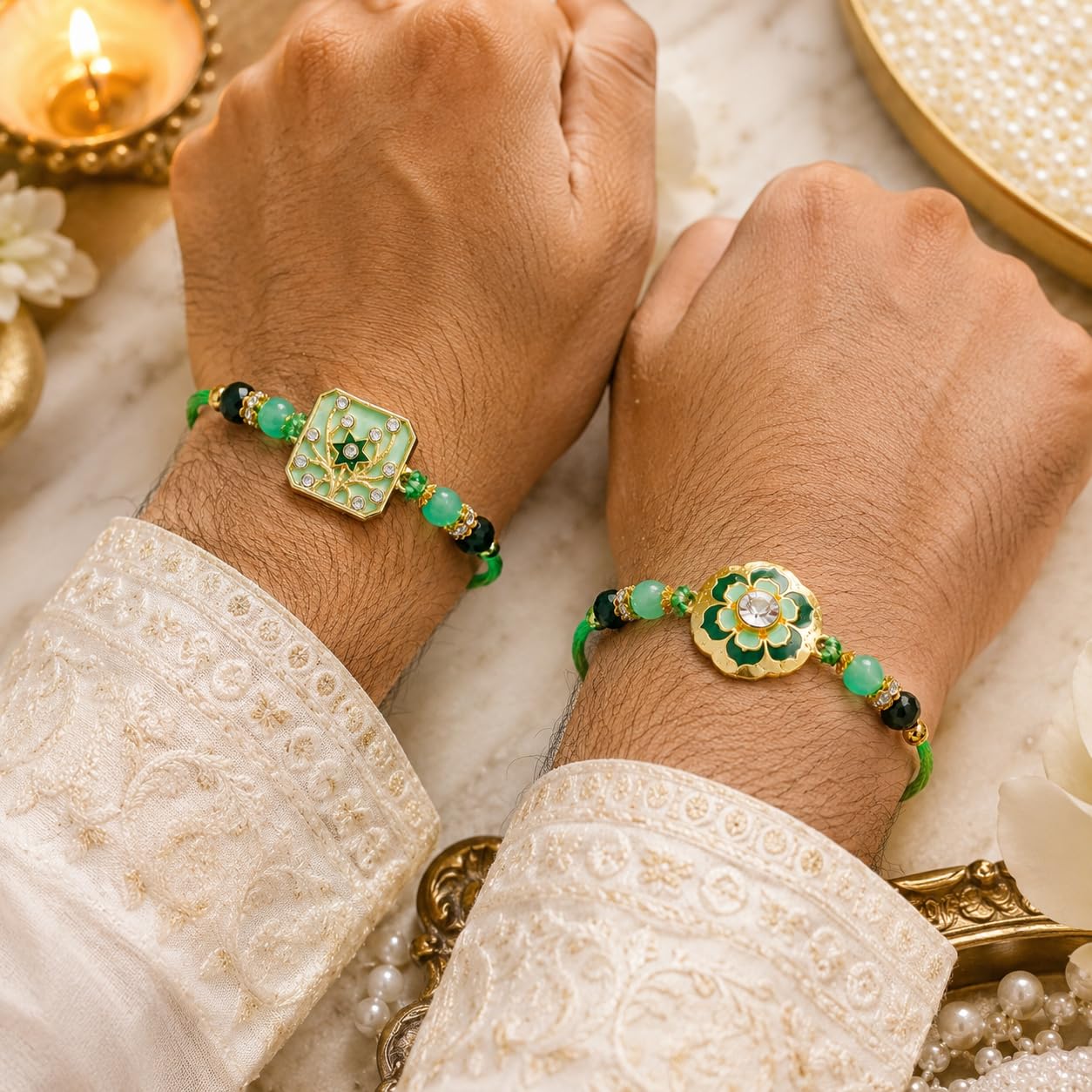 Two hands wearing matching green and gold beaded bracelets with decorative floral and square charms, shown against a warm marble and lace background with a lit candle.