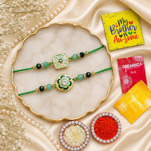 Two green thread Rakhi bracelets with gold and green floral motifs and beads are arranged on a scalloped cream tray. The scene includes pearl strands, dried flowers, small bowls of rice and red powder, and two branded packets reading 'My Brother Is Awesome' and 'DREAMICA Roll' and 'DREAMICA Chai'.