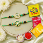 Two decorative Rakhi bracelets with green cords, turquoise beads, and ornate gold-toned floral and geometric motifs are arranged on a beige oval tray. The scene includes white flowers, pearl-like beads, a yellow card reading 'My Brother is Awesome,' and packets of DREAMICA roli and chana powder.