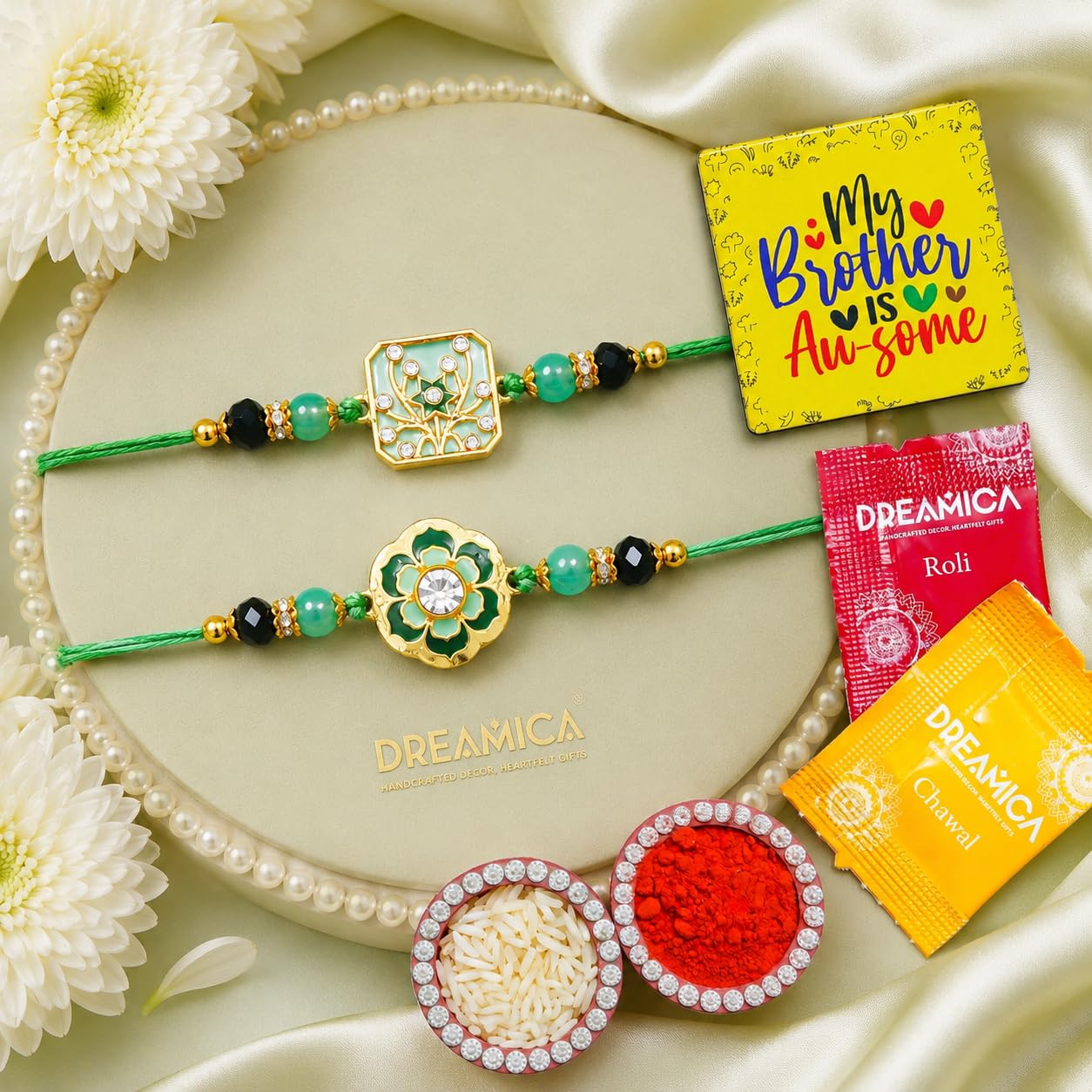 Two decorative Rakhi bracelets with green cords, turquoise beads, and ornate gold-toned floral and geometric motifs are arranged on a beige oval tray. The scene includes white flowers, pearl-like beads, a yellow card reading 'My Brother is Awesome,' and packets of DREAMICA roli and chana powder.