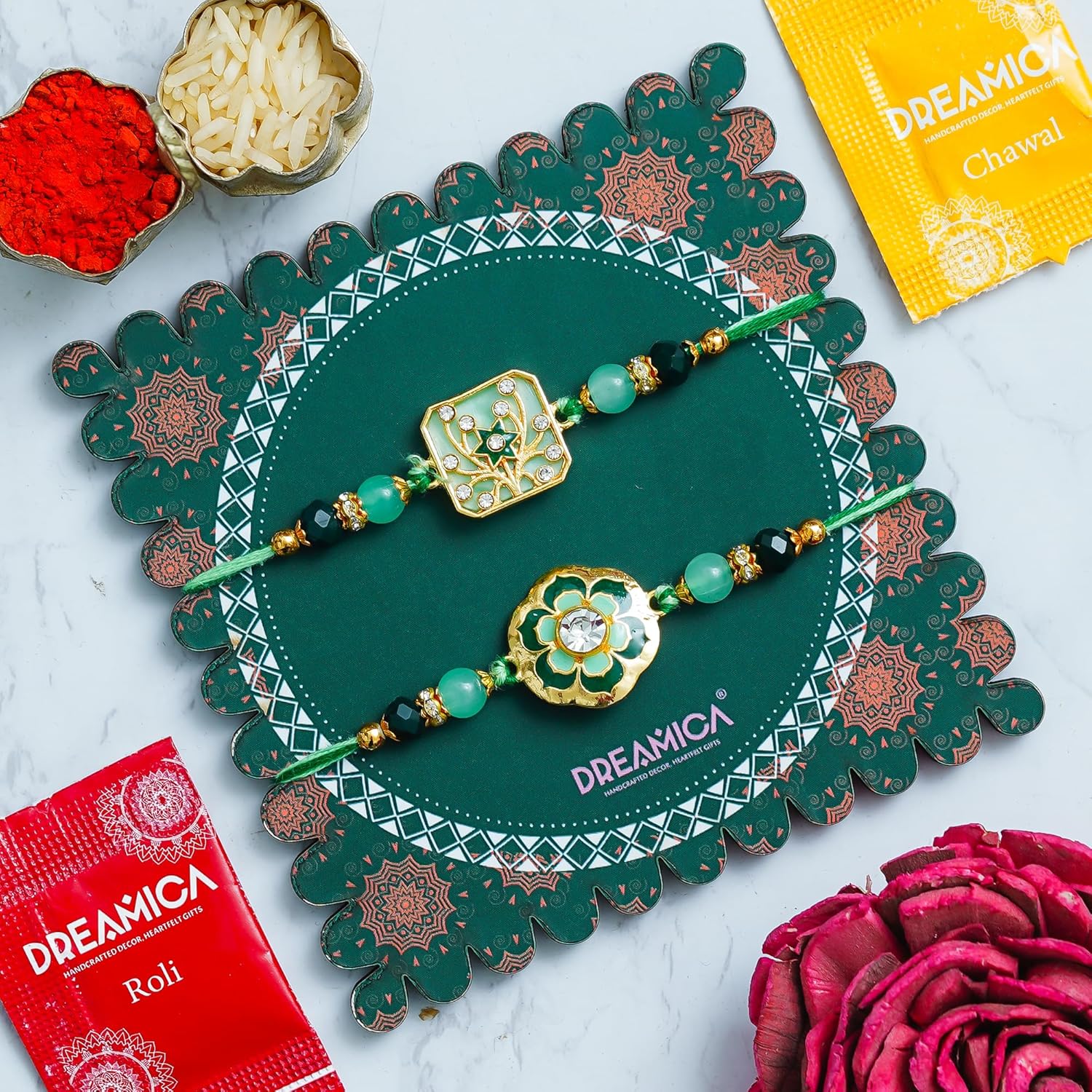 Two decorative Rakhi bracelets displayed on a green scalloped card, surrounded by red and yellow Dreamica packaging and small bowls of ceremonial items on a light marble surface.